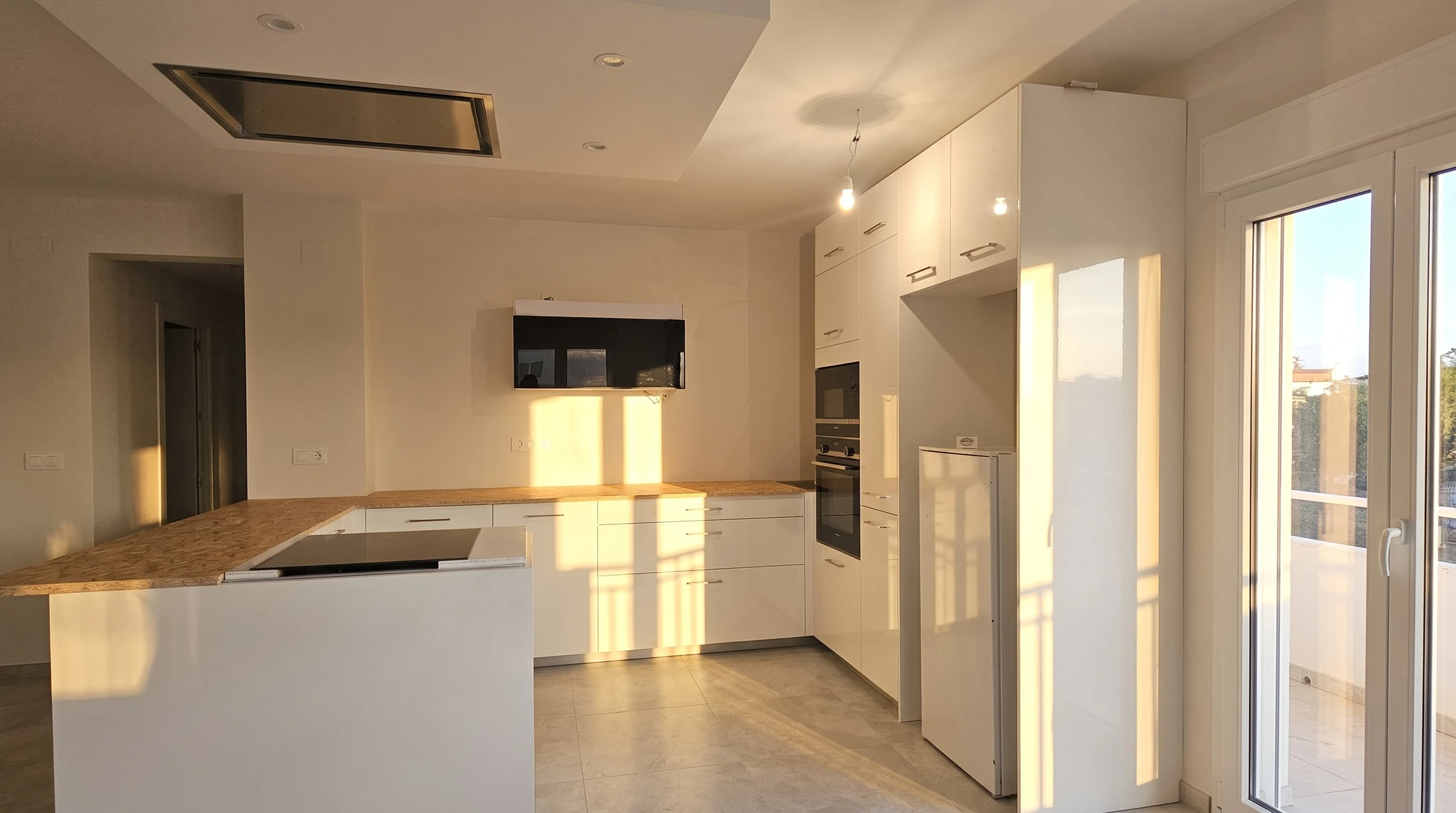 Photographie d'un intérieur résidentiel rénové de manière complète : grande pièce ouverte combinant cuisine moderne et séjour. Au premier plan, une cuisine équipée avec meubles, plan de travail, plaque de cuisson et électroménagers. Au second plan, un séjour avec canapé, murs fraîchement peints, menuiseries neuves, sol en parquet harmonisé. Éclairage moderne intégré, crédence et accessoires de finition de qualité. La scène doit clairement montrer une transformation globale d'espace, illustrant la coordination de multiples corps de métier dans un même projet. Ambiance professionnelle, chaleureuse et aspirationnelle.